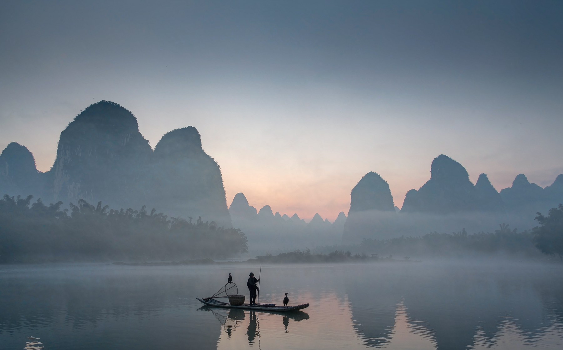 Li River at dawn near Guilin, China - layered karst peaks rising through morning mist with a bamboo raft and cormorant fisherman on still water